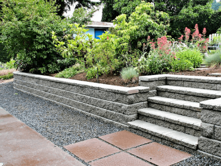 Textured grey stone retaining wall and stairs bordering a lush garden with pink flowers.