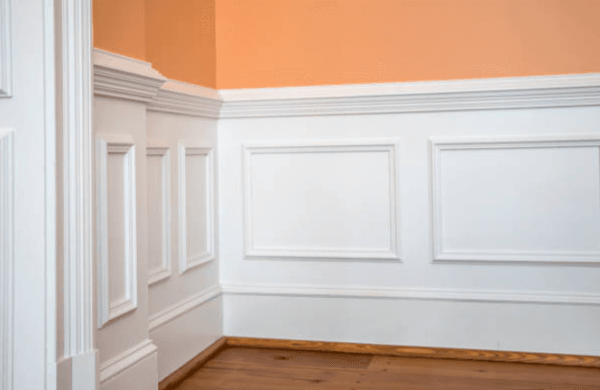 White raised panel wainscoting below a peach wall with dark hardwood floors in a corner.